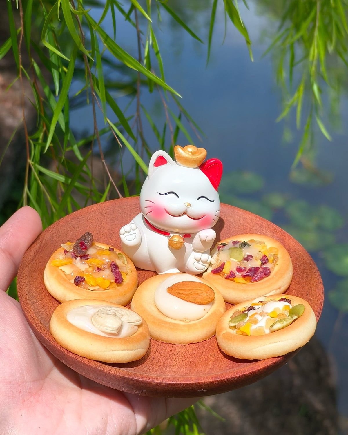 Hand holding a small wooden plate with mini pizzas and a cat figurine against a natural background