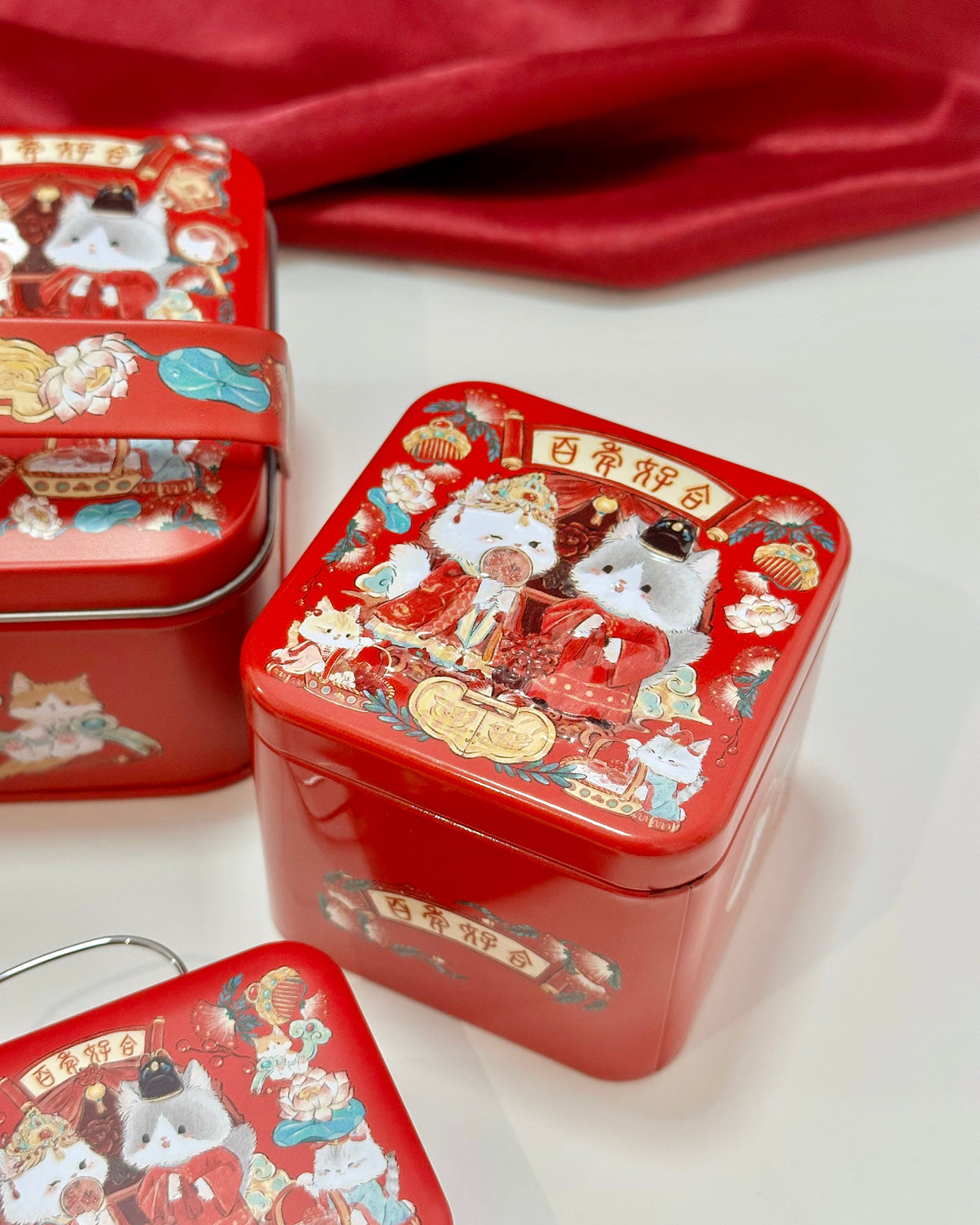 Three red decorative metal gift tins with illustrated cat characters and Chinese wedding style motifs, arranged on a white surface with a draped red fabric in the background.
