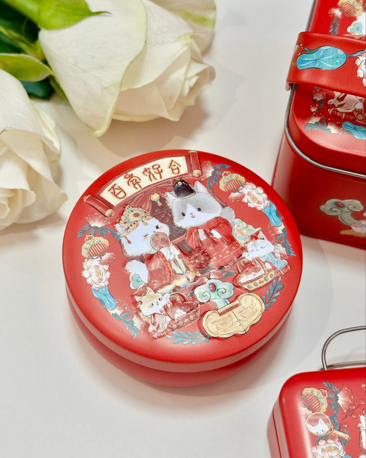 A round red wedding decorative tin with a festive illustrated lid featuring cute cartoon animals cats and Chinese traditional wedding motifs, styled on a white surface with white roses and additional matching red tins partially visible.