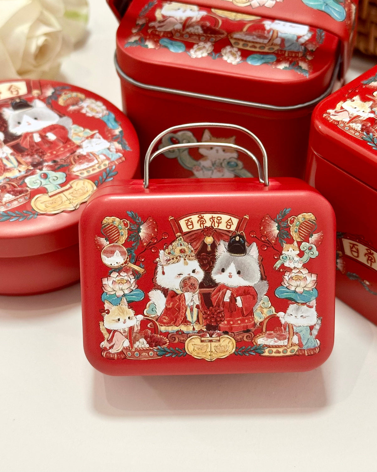 A red metal gift tin with a handle in the foreground, surrounded by similar red tins. The tins are decorated with colorful cartoon cat illustrations and Chinese traditional wedding themed graphics, including gold Chinese characters and festive motifs.