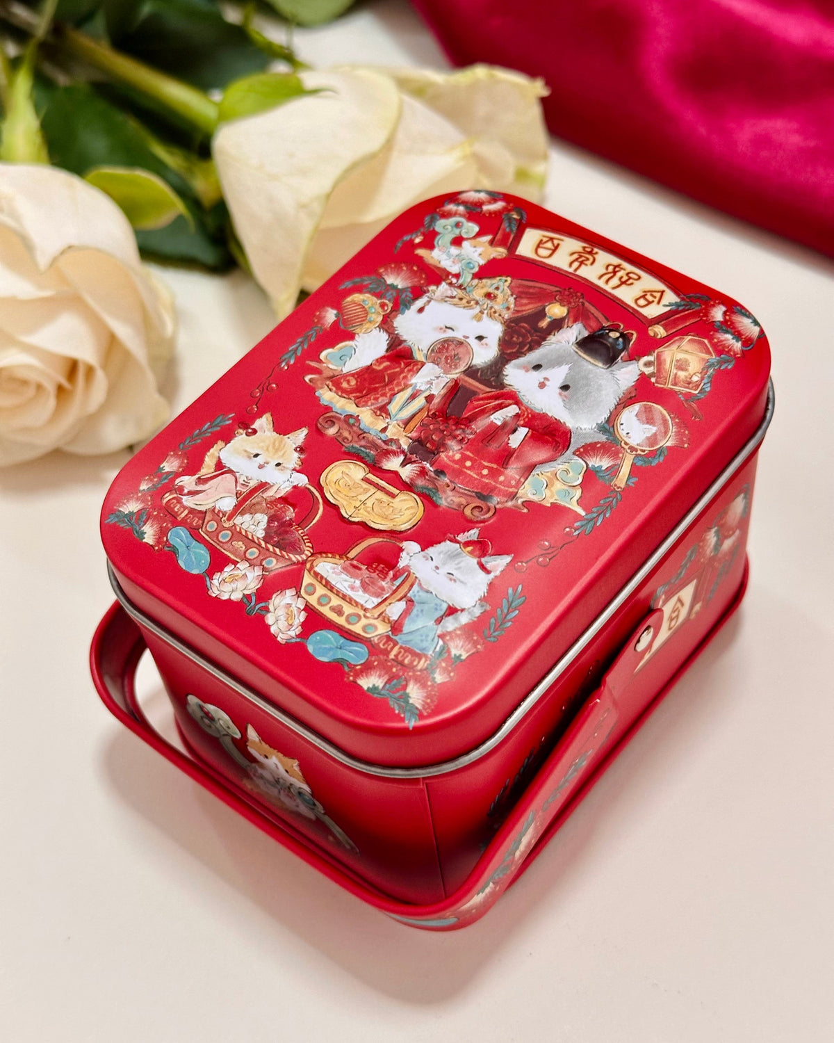 A red rectangular metal tin with a handle, decorated with wedding cute cartoon cat illustrations and Chinese traditional Wedding style, shown on a light surface with cream roses and a red fabric in the background.