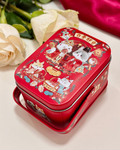 A red rectangular metal tin with a handle, decorated with wedding cute cartoon cat illustrations and Chinese traditional Wedding style, shown on a light surface with cream roses and a red fabric in the background.