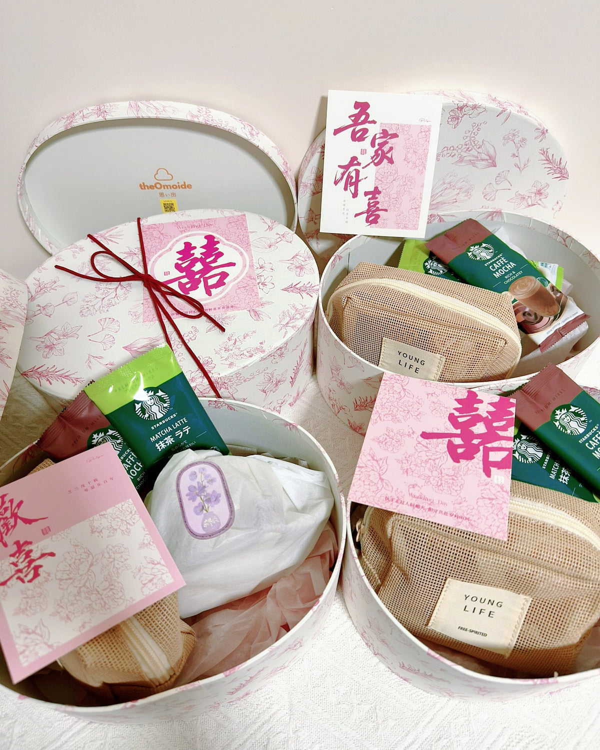 Three round gift boxes filled with assorted packaged items, including a white pouch, beige mesh pouches, tea packets, and pink cards with Chinese characters, arranged on a light fabric surface.