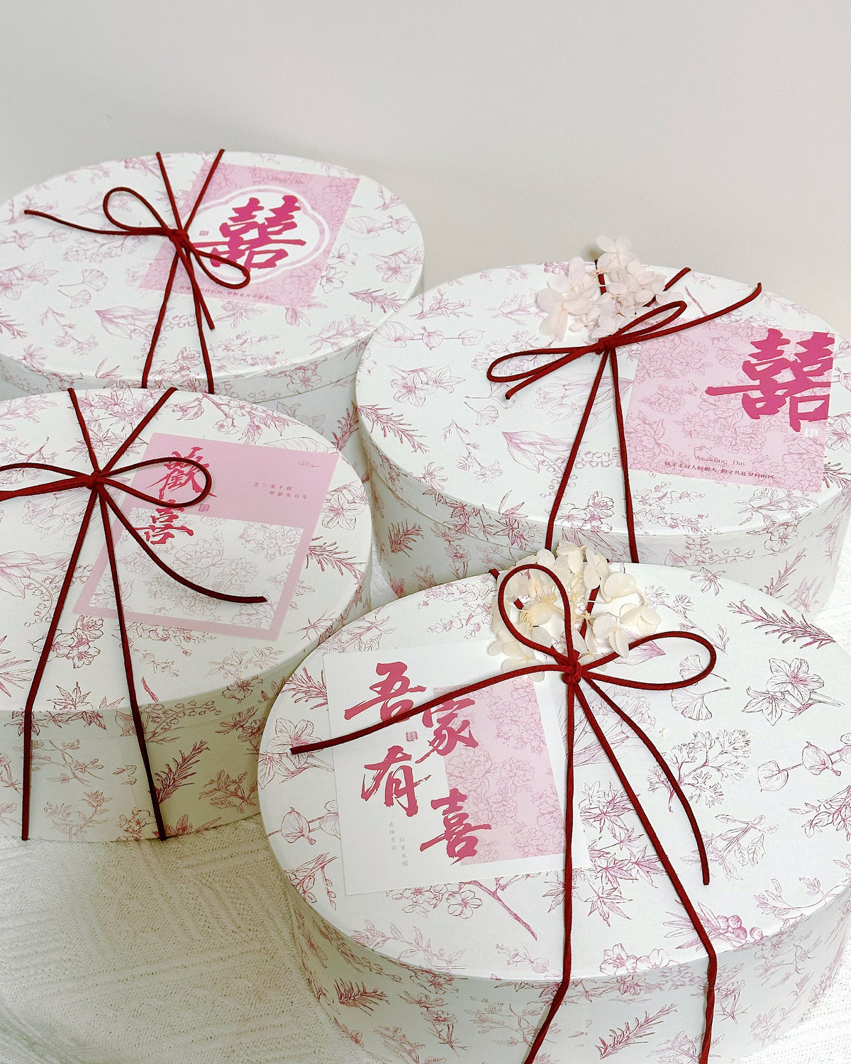 Four round gift boxes with white floral-patterned lids, tied with dark red ribbon bows and decorated with pink Chinese characters, arranged on a white surface against a light background.