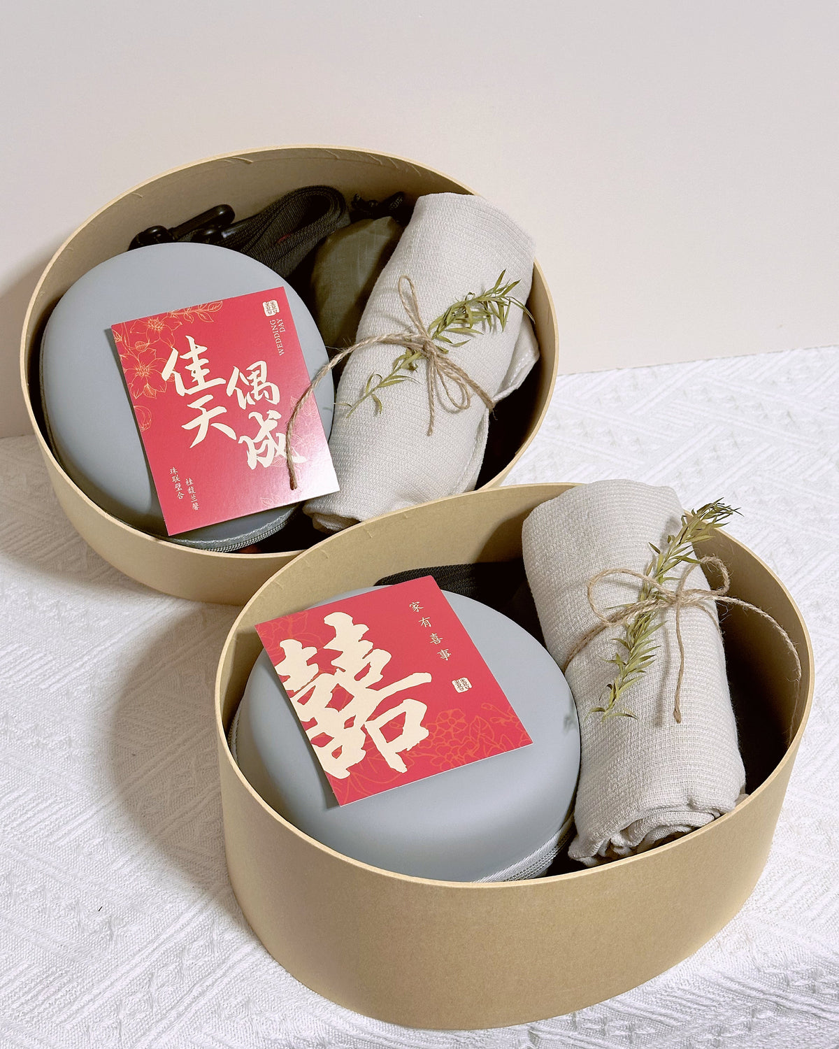Two round beige gift boxes on a white textured surface, each containing a round gray item with a red card featuring Chinese characters, alongside a rolled cream fabric tied with twine and a small sprig of greenery.