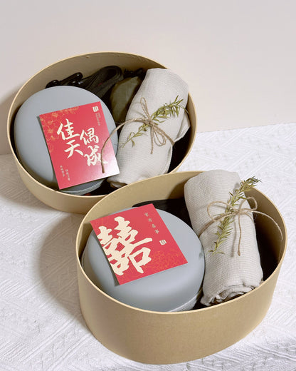 Two round beige gift boxes on a white textured surface, each containing a round gray item with a red card featuring Chinese characters, alongside a rolled cream fabric tied with twine and a small sprig of greenery.