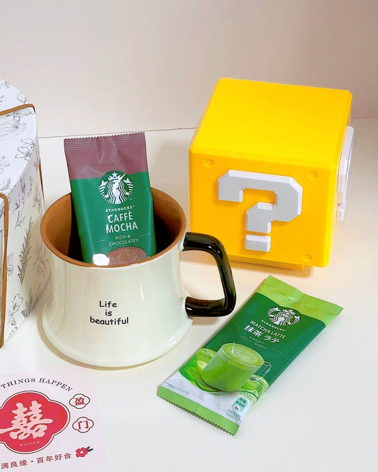 A styled tabletop scene with a white ceramic mug labeled 'Life is beautiful' containing a green Starbucks Caffe Mocha packet, a yellow block with a white question mark, another green Starbucks packet, and part of a patterned box on a light background.