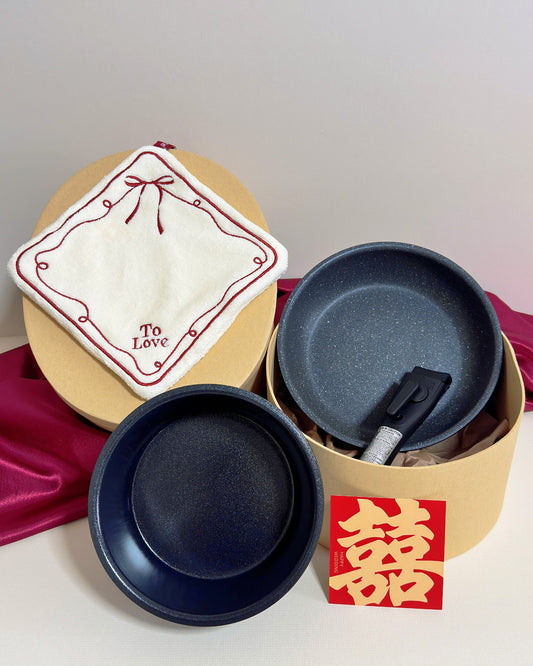 A set of three round shallow pans in different sizes, with one pan showing a dark gray exterior and another showing a dark navy interior. A white embroidered cloth hand towel with red trim and the words 'To Love' is draped over the largest pan, which sits inside a kraft paper round box.