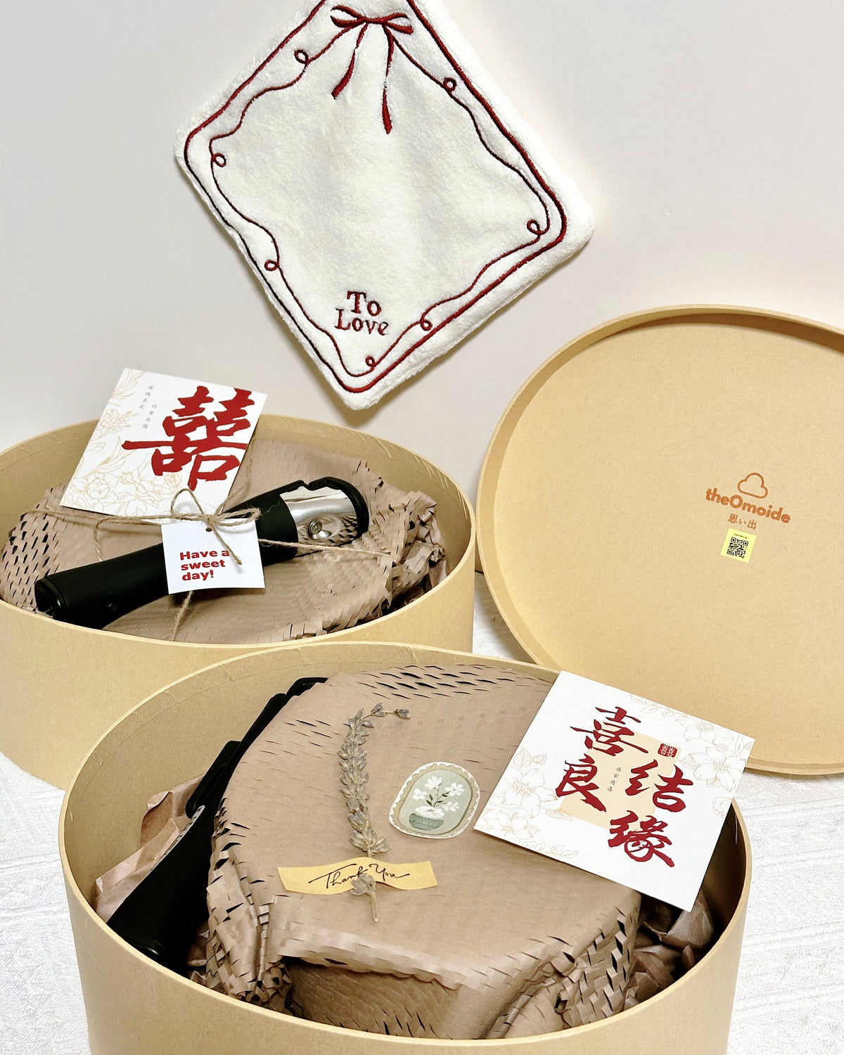 A wedding gift set presented in round beige boxes with the lid off, showing a cream embroidered 'To Love' hand towel, a black-and-silver pocket pan or pot handle, a beige woven textile with a decorative tag, and a small card with Chinese characters. The brand name 'theOrnside' is visible on the lid.