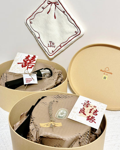 A wedding gift set presented in round beige boxes with the lid off, showing a cream embroidered 'To Love' hand towel, a black-and-silver pocket pan or pot handle, a beige woven textile with a decorative tag, and a small card with Chinese characters. The brand name 'theOrnside' is visible on the lid.