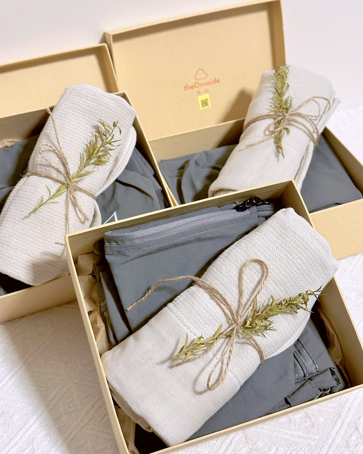 Three beige muji style travel gift boxes containing folded gray travel organizers and rolled cream towels tied with twine and a small dried green sprig, arranged on a white textured surface.