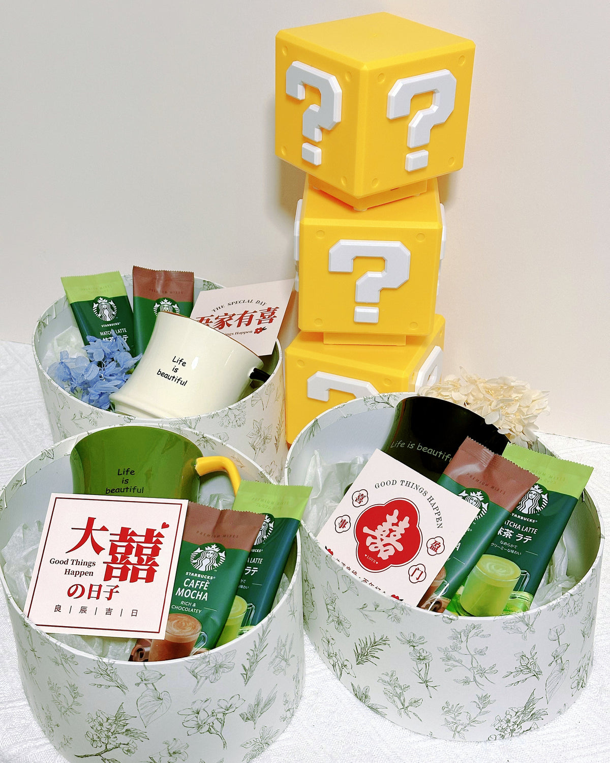 A gift basket set featuring three white patterned cylindrical boxes filled with assorted packaged snacks, drinks, and small items, arranged with three stacked yellow question-block cubes on a white surface against a light background.