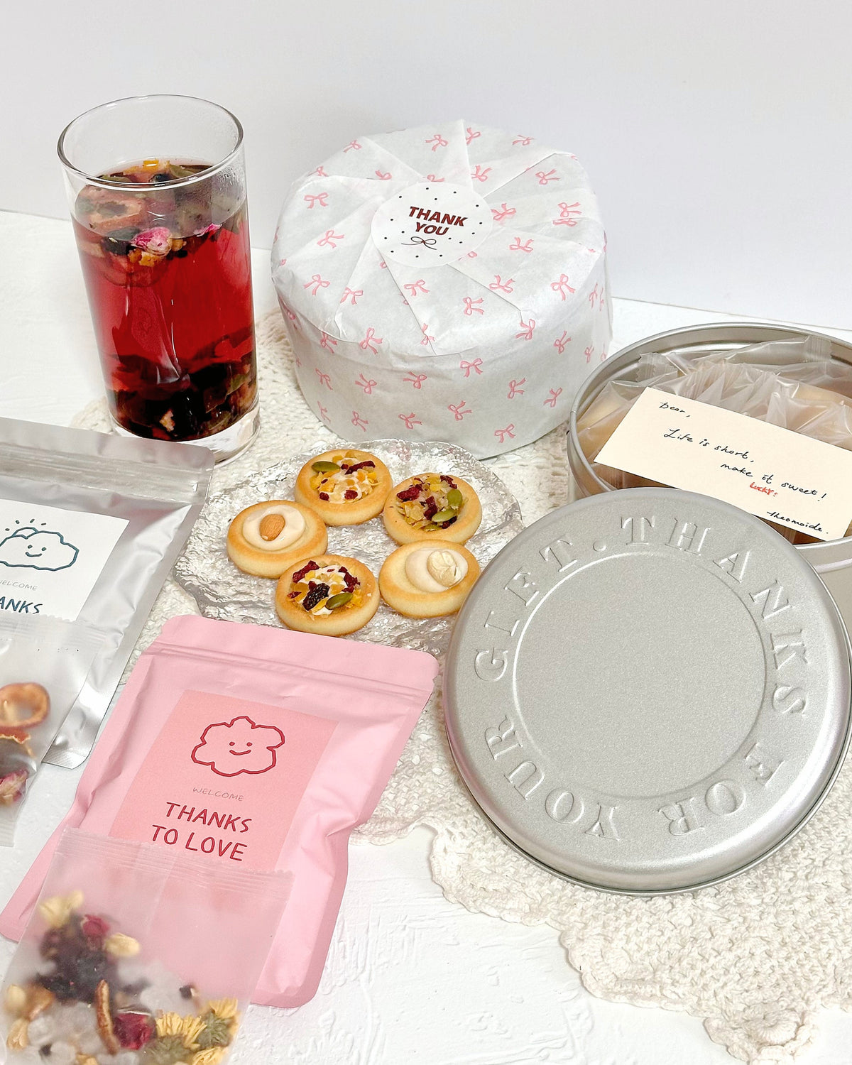 A gift set arranged on a white surface, including a pink pouch labeled 'THANKS TO LOVE,' a glass of red tea, small cookies, a round wrapped item with 'THANKS YOU!' text, a metal tin lid, and a clear pouch with dried flower pieces.
