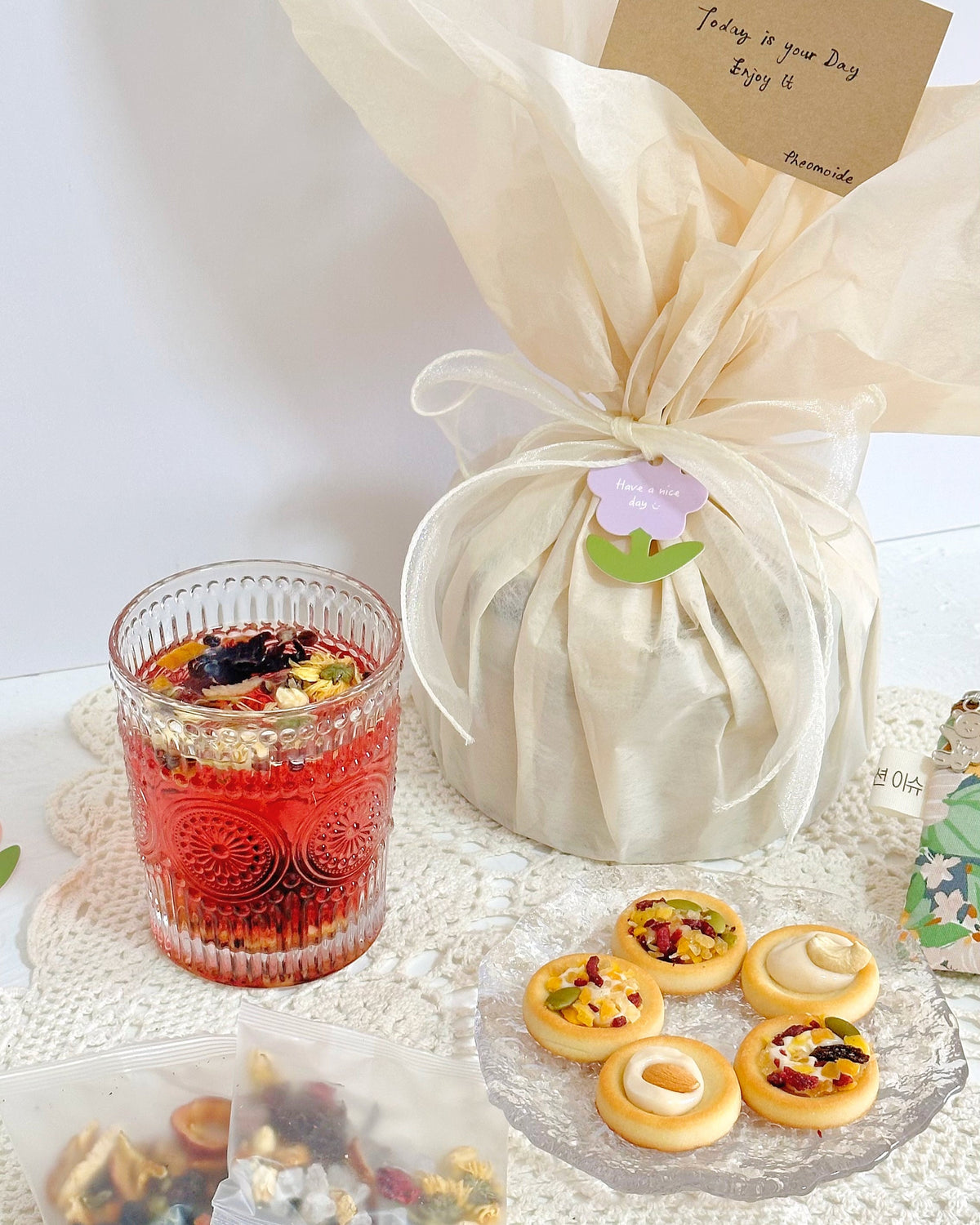 A gift-wrapped bundle tied with a ribbon and small flower charm, alongside a glass of red herbal tea and several small pastries on a white lace doily.
