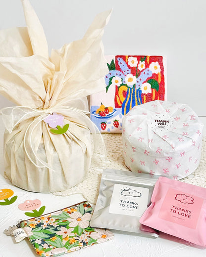 A gift set arranged on a white surface with a cream tissue-wrapped box as the main subject, alongside a pink floral pouch, a silver pouch, a pink 'THANKS TO LOVE' card, a small floral card, and a colorful butterfly artwork in the background.