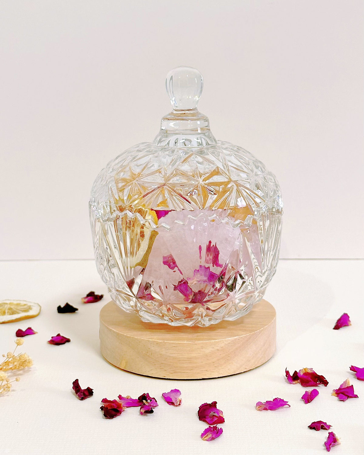 A clear cut-glass jar with a matching glass lid sits on a round light wood base, containing dried pink flower petals and botanical pieces. The setup is photographed on a white surface with scattered pink petals around it against a plain white background.