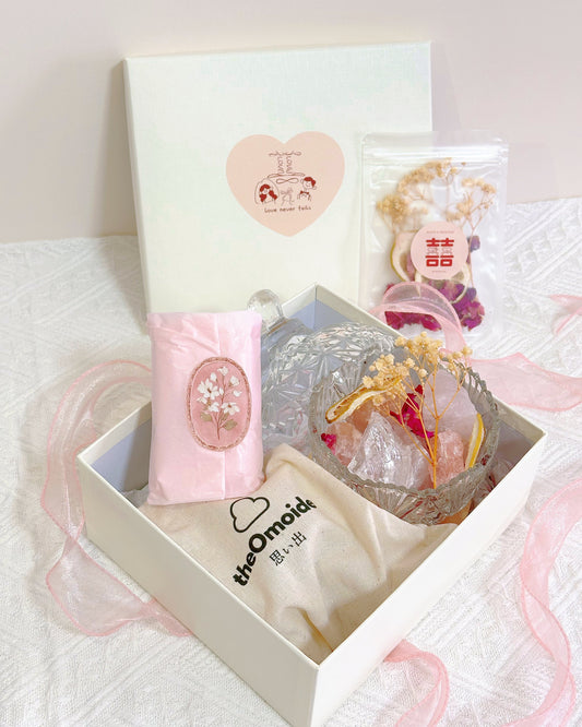 A gift box set containing a pink sachet aroma essential oil, a clear glass bowl with dried roses flowers, pink healing romance crystal, a cream-colored pouch printed with 'the Omoide, and a small clear bag with a red double happiness symbol, arranged in a white box on a white textured fabric background