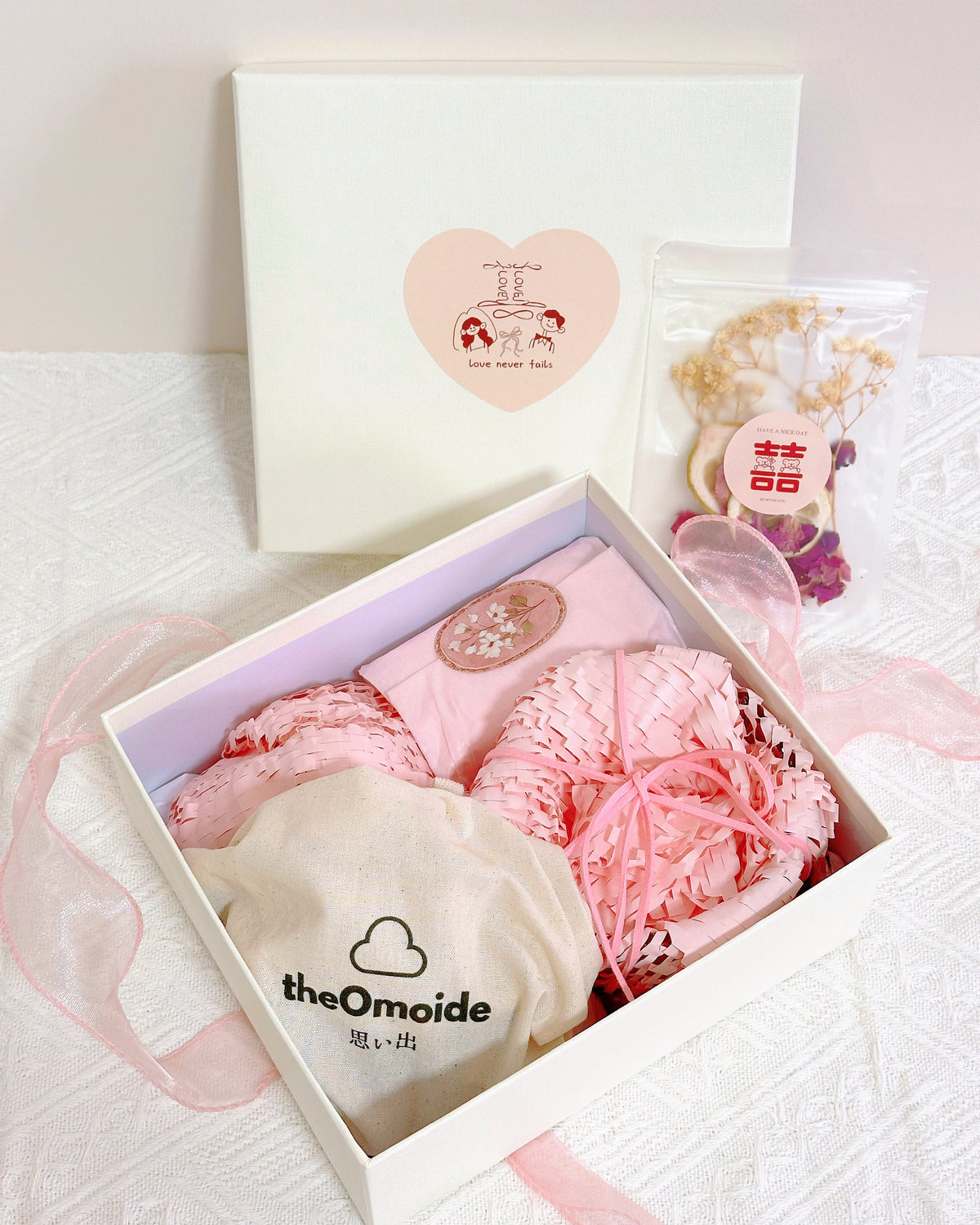 A pink baby gift set in a white box, featuring a pink textured wrap, a cream fabric pouch printed with 'theOmoide', and a small clear bag of dried flowers, arranged on a white textured surface with a white gift box lid in the background.
