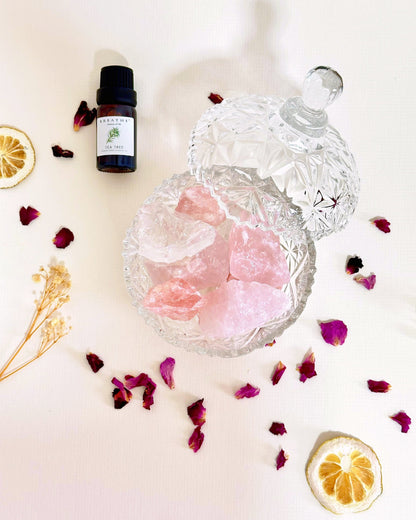 A styled flat-lay featuring a small amber dropper bottle labeled 'Balm of Nature' next to a clear cut-glass jar filled with pink bath salts, surrounded by dried rose petals, dried lemon slices, and a sprig of wheat on a white background.
