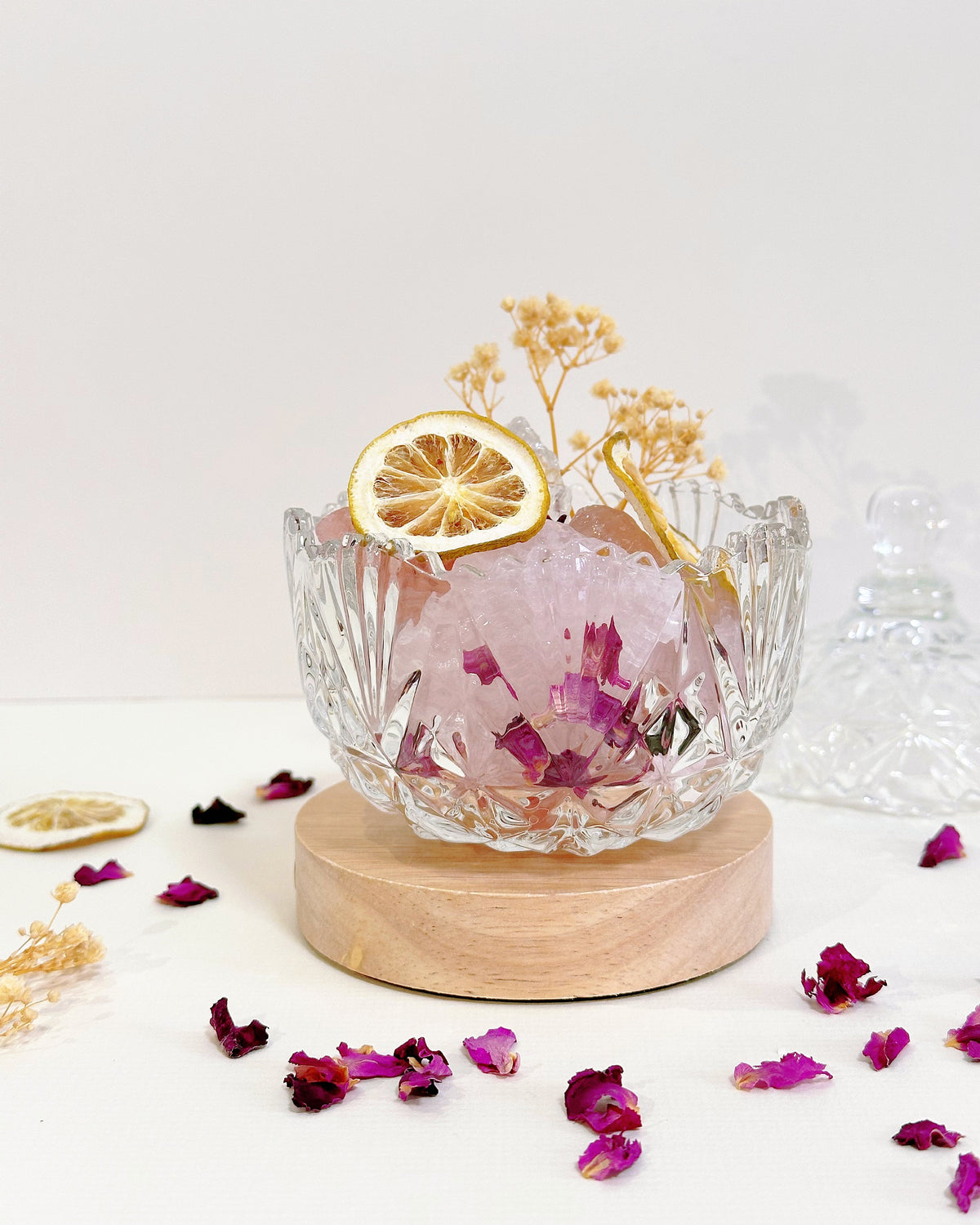 A clear glass bowl filled with pink bath salts and dried flower petals, styled on a round wooden coaster with a dried orange slice garnish, against a clean white background.