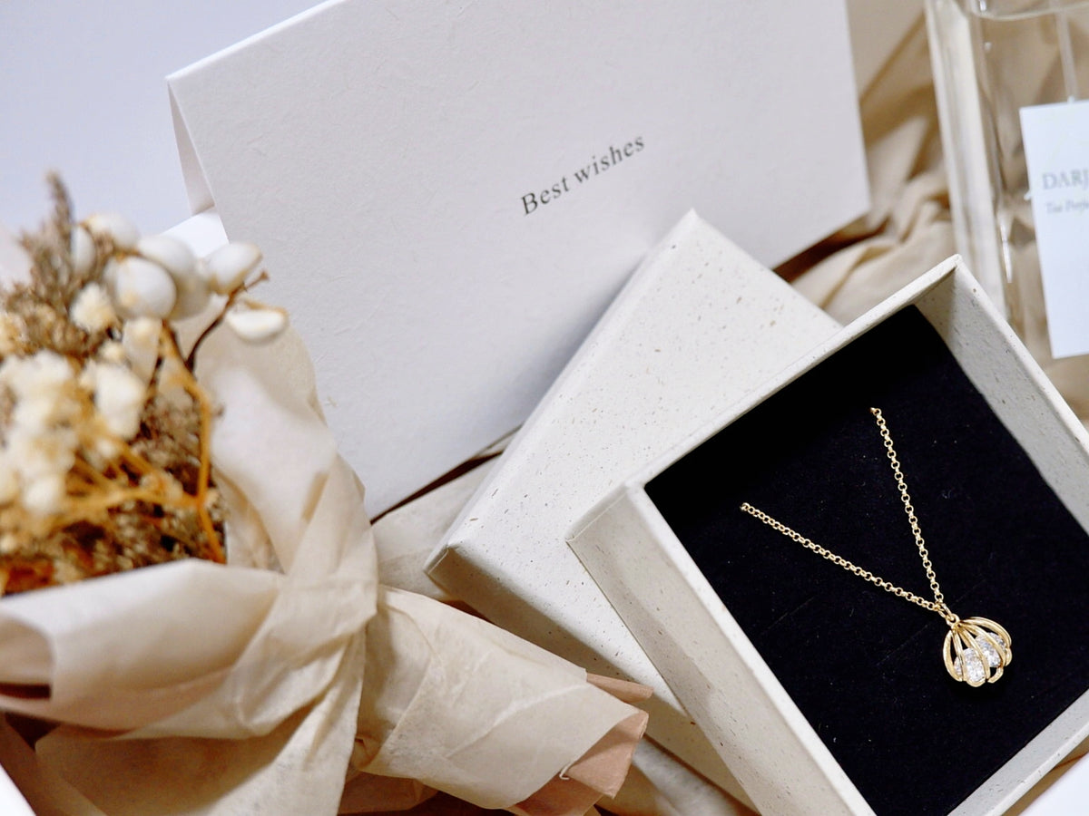 A gold pendant necklace with clear stones displayed in an open white jewelry box with a black velvet insert. A closed white box with the words 'Best wishes' is behind it, alongside cream-colored ribbon and dried floral decor.