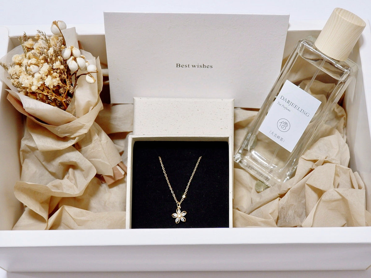 A gift box presentation featuring a gold flower pendant necklace in a black box, a bottle of Darjeeling tea with a cream cap, dried flowers, and beige tissue paper inside a white box.