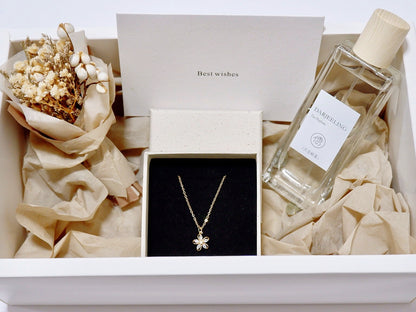 A gift box presentation featuring a gold flower pendant necklace in a black box, a bottle of Darjeeling tea with a cream cap, dried flowers, and beige tissue paper inside a white box.