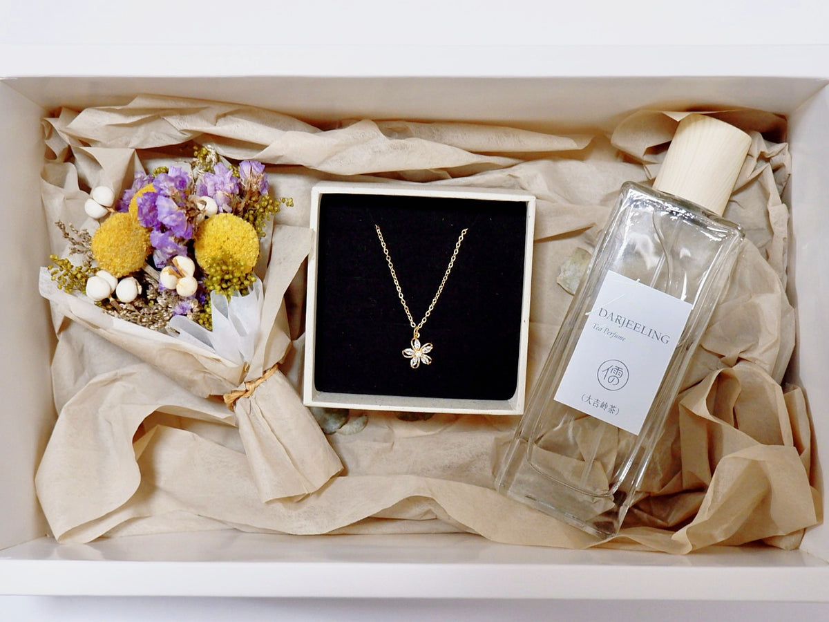 A gift box arrangement featuring a delicate gold flower pendant necklace on a black display card, a clear glass perfume bottle labeled 'DUBLING' with a cream cap, and a small bouquet of dried flowers, all nestled in beige tissue paper.
