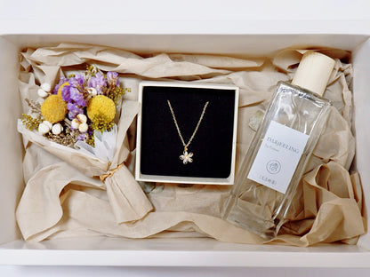 A gift box arrangement featuring a delicate gold flower pendant necklace on a black display card, a clear glass perfume bottle labeled 'DUBLING' with a cream cap, and a small bouquet of dried flowers, all nestled in beige tissue paper.