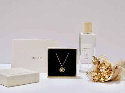 A gold necklace with a shell-shaped pendant displayed in a black box, alongside a cream-colored gift box, a clear glass perfume bottle labeled 'Tea Perfume', and dried baby's breath flowers magnet on a white surface.