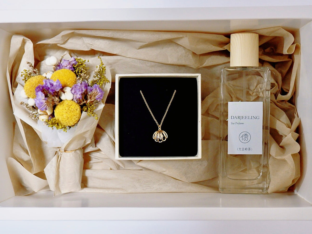 A gift box arrangement featuring a gold pendant necklace in a black box, a small bouquet of yellow and purple dried flowers, and a clear glass perfume bottle labeled 'DARFEELING Eau Parfum' on beige tissue paper.