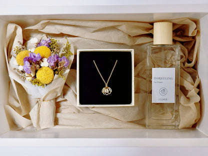 A gift box arrangement featuring a gold pendant necklace in a black box, a small bouquet of yellow and purple dried flowers, and a clear glass perfume bottle labeled 'DARFEELING Eau Parfum' on beige tissue paper.