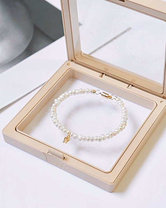 A pearl beaded bracelet with a small gold charm, displayed inside an open beige jewelry box with a clear lid.