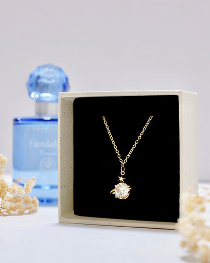 A gold pendant necklace displayed inside an open cream-colored jewelry box with a black interior. The pendant appears to be a small round clear stone in a gold setting, suspended from a delicate gold chain.