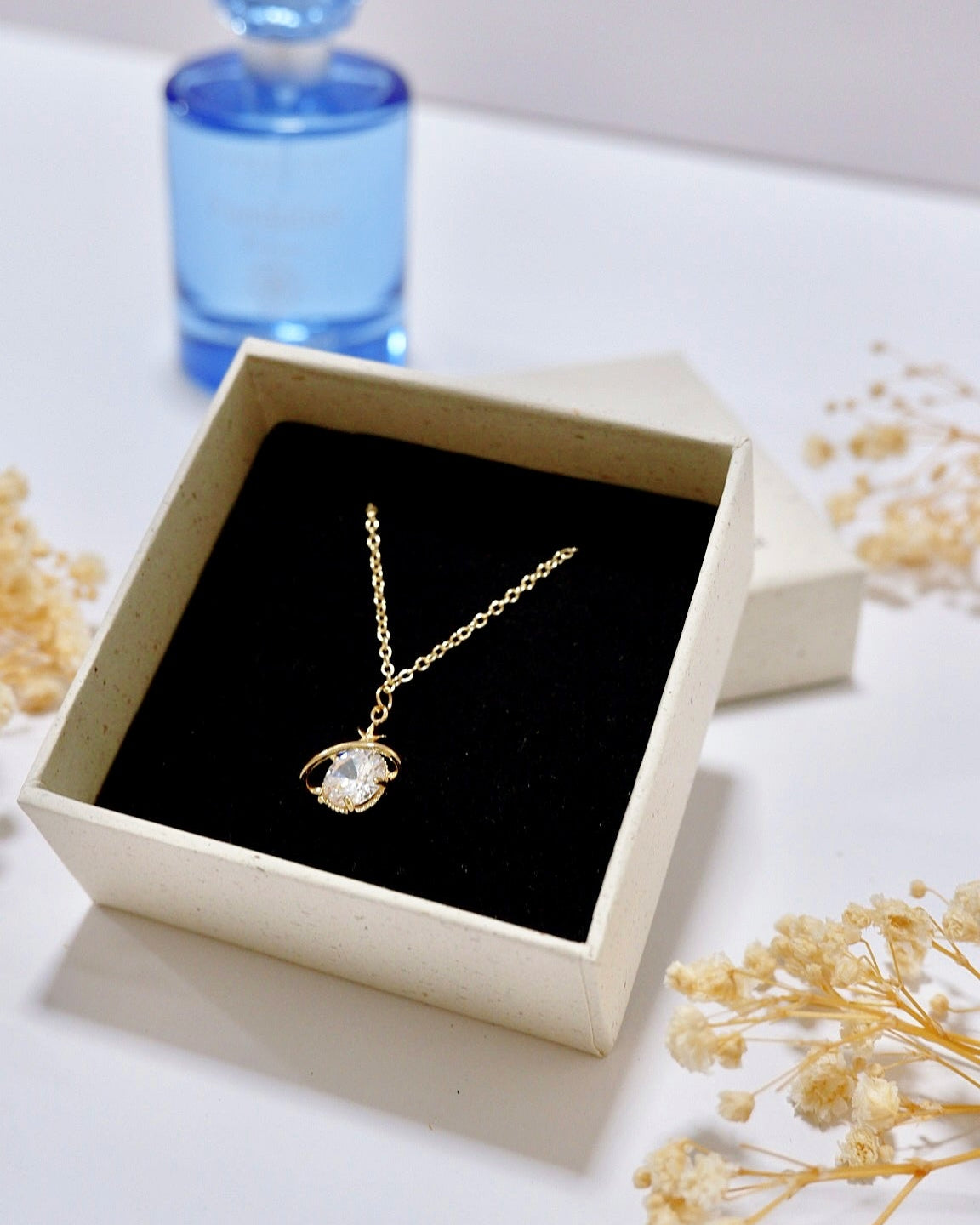A gold pendant necklace with a clear round gemstone displayed in an open cream-colored jewelry box with a black velvet insert. A blue perfume bottle is blurred in the background on a white surface, with dried flowers scattered around.