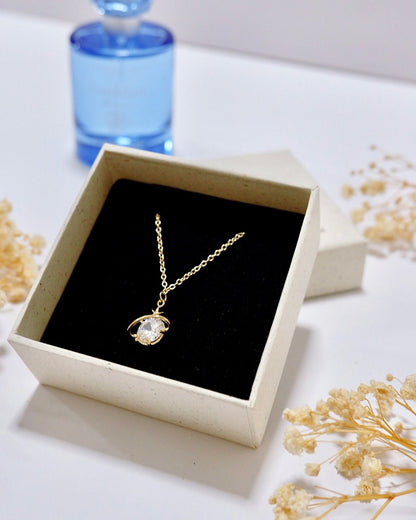 A gold pendant necklace with a clear round gemstone displayed in an open cream-colored jewelry box with a black velvet insert. A blue perfume bottle is blurred in the background on a white surface, with dried flowers scattered around.