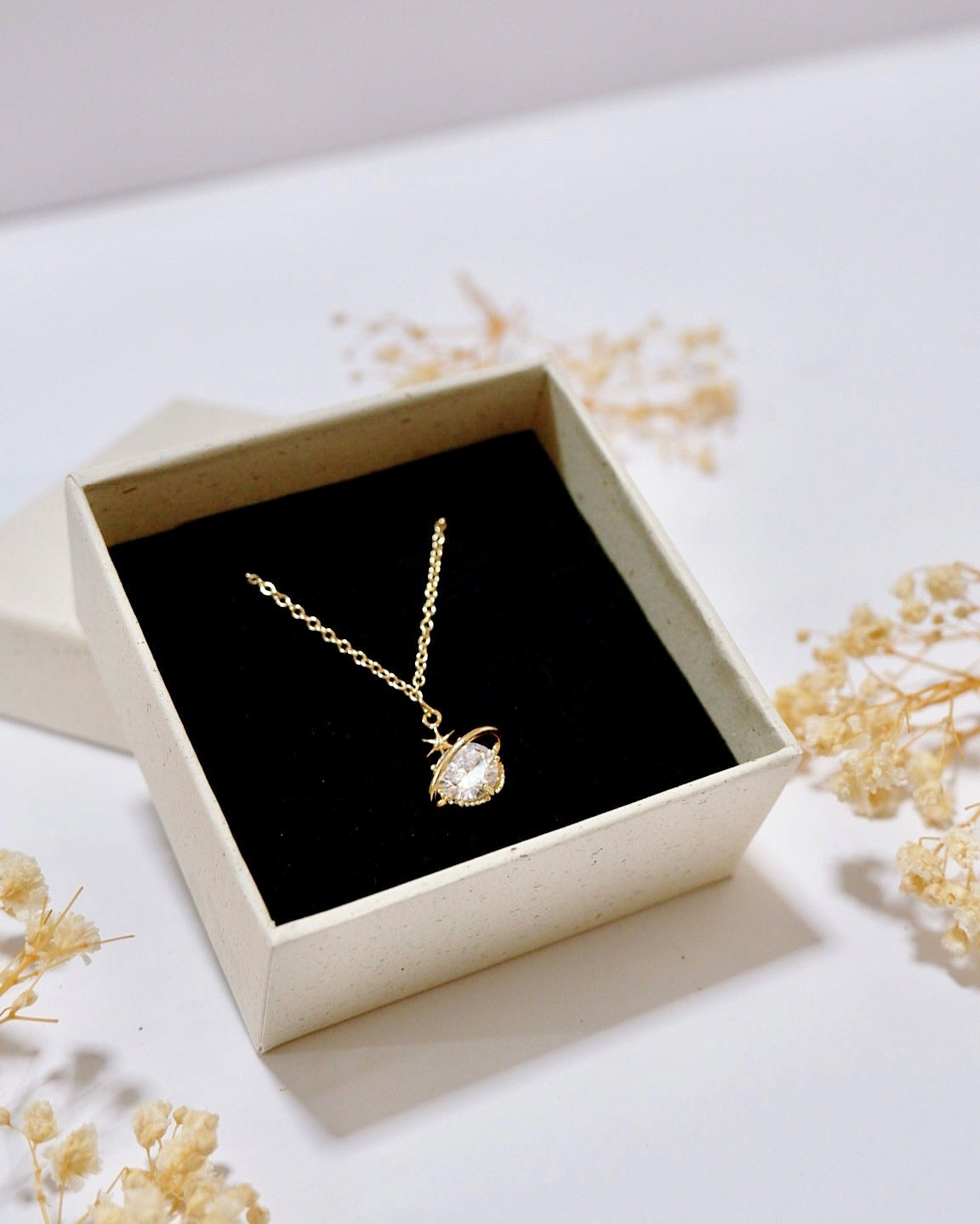 A gold pendant necklace with a clear teardrop-shaped gemstone displayed inside an open cream-colored gift box with a black velvet lining, surrounded by dried baby's breath flowers on a white surface.