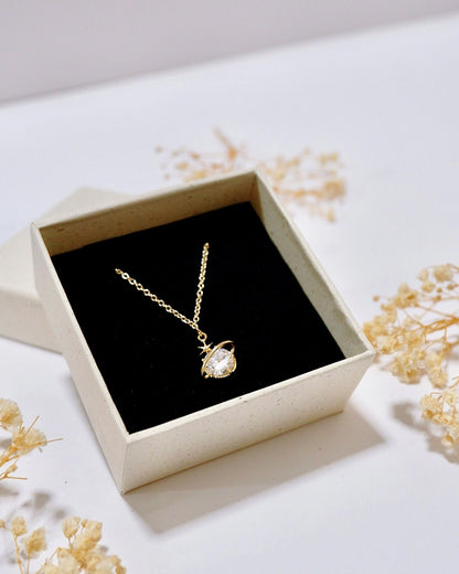 A gold pendant necklace with a clear teardrop-shaped gemstone displayed inside an open cream-colored gift box with a black velvet lining, surrounded by dried baby's breath flowers on a white surface.