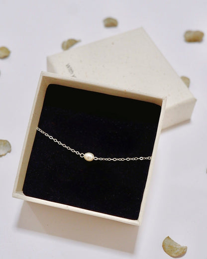 A delicate silver chain bracelet with a single white pearl bead, presented in an open cream-colored gift box with a black velvet lining, set against a white background with scattered decorative petals.