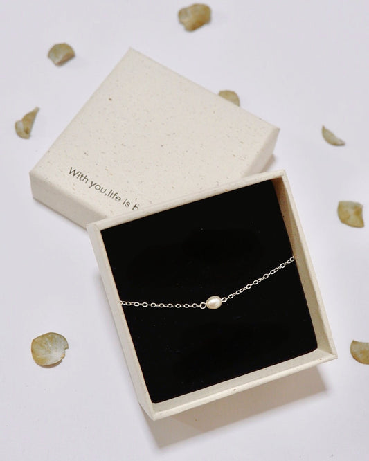A delicate silver chain bracelet with a single small pearl bead, presented in an open cream-colored gift box with a black interior. The box lid is placed behind it on a white surface scattered with small beige flower petals.