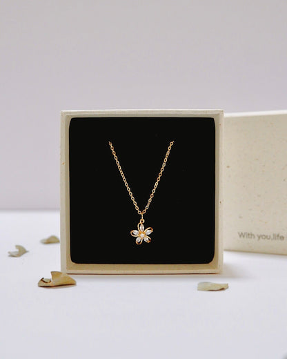 A delicate gold chain necklace with a small flower-shaped pendant displayed inside an open cream-colored jewelry box with a black insert, set against a clean white background.
