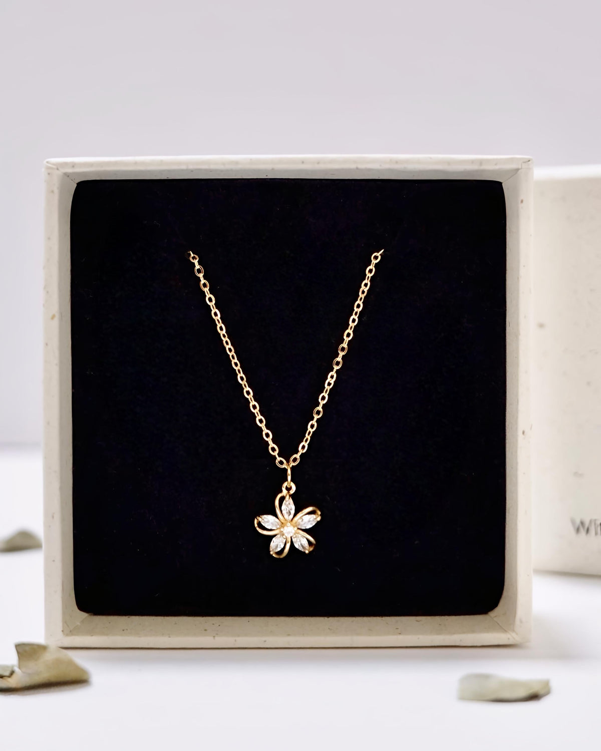 A delicate gold chain necklace with a small flower-shaped pendant, presented inside a square black velvet jewelry box with a cream-colored frame.