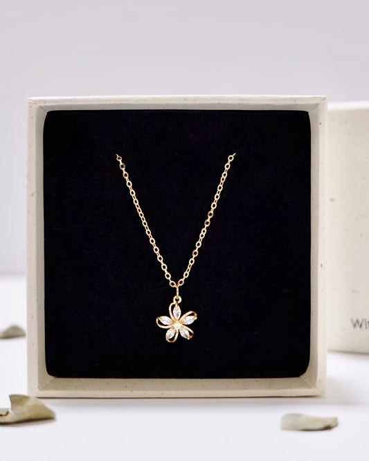 A delicate gold chain necklace with a small flower-shaped pendant, presented inside a square black velvet jewelry box with a cream-colored frame.
