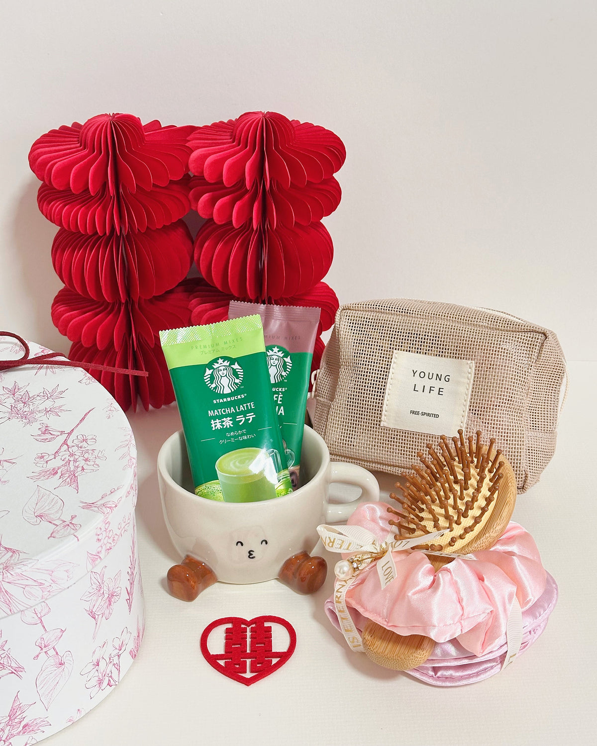 A curated gift set featuring a red paper honeycomb box, a Starbucks Matcha Latte pouch, a beige canvas pouch labeled 'YOUNG LIFE', a pink satin hair scrunchie with wooden hairbrushes, a white mug with a cartoon face, and a red heart-shaped ornament on a white background.