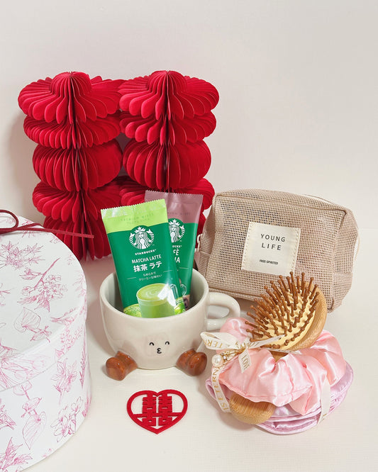 A curated gift set featuring a red paper honeycomb box, a Starbucks Matcha Latte pouch, a beige canvas pouch labeled 'YOUNG LIFE', a pink satin hair scrunchie with wooden hairbrushes, a white mug with a cartoon face, and a red heart-shaped ornament on a white background.