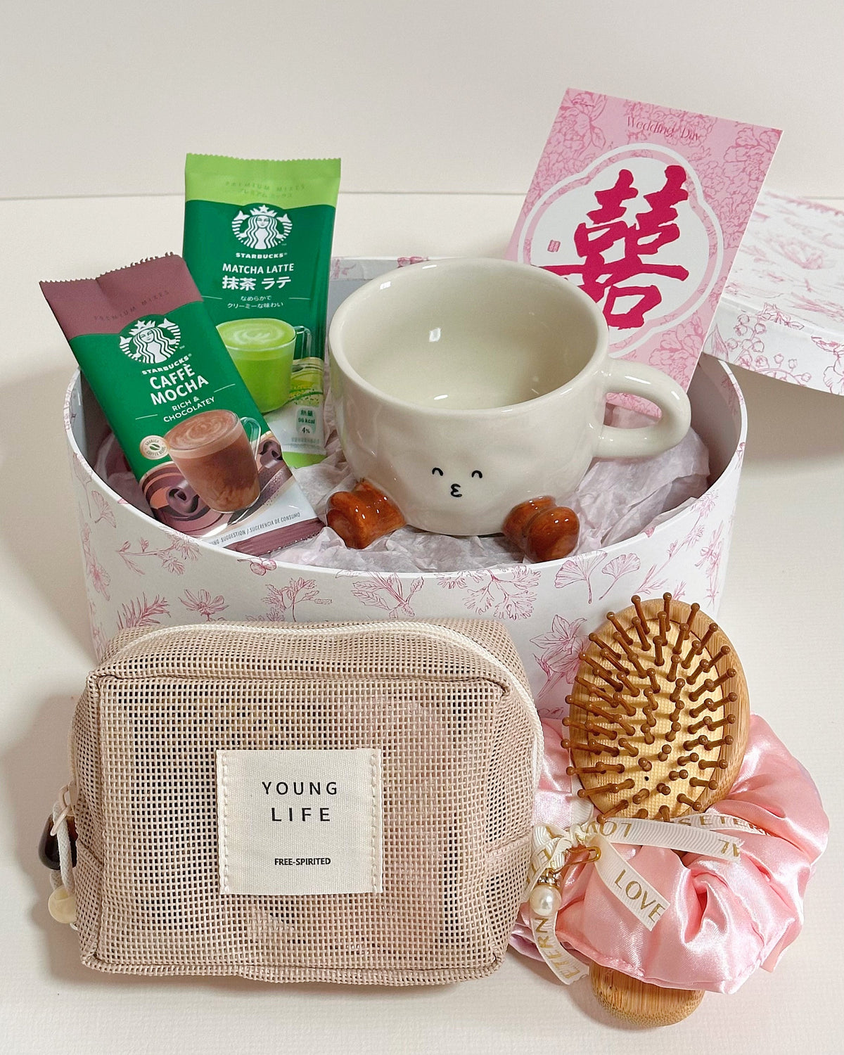 A curated gift set arranged in a round pink patterned box on a white surface. The set includes a white ceramic mug with a cute face, two Starbucks coffee pouches (caramel mocha and green tea latte), a pink card with a red Chinese character, a beige mesh pouch labeled 'YOUNG LIFE', and a wooden hairbrush tied with a pink satin ribbon.