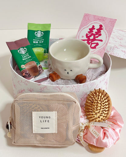 A curated gift set arranged in a round pink patterned box on a white surface. The set includes a white ceramic mug with a cute face, two Starbucks coffee pouches (caramel mocha and green tea latte), a pink card with a red Chinese character, a beige mesh pouch labeled 'YOUNG LIFE', and a wooden hairbrush tied with a pink satin ribbon.