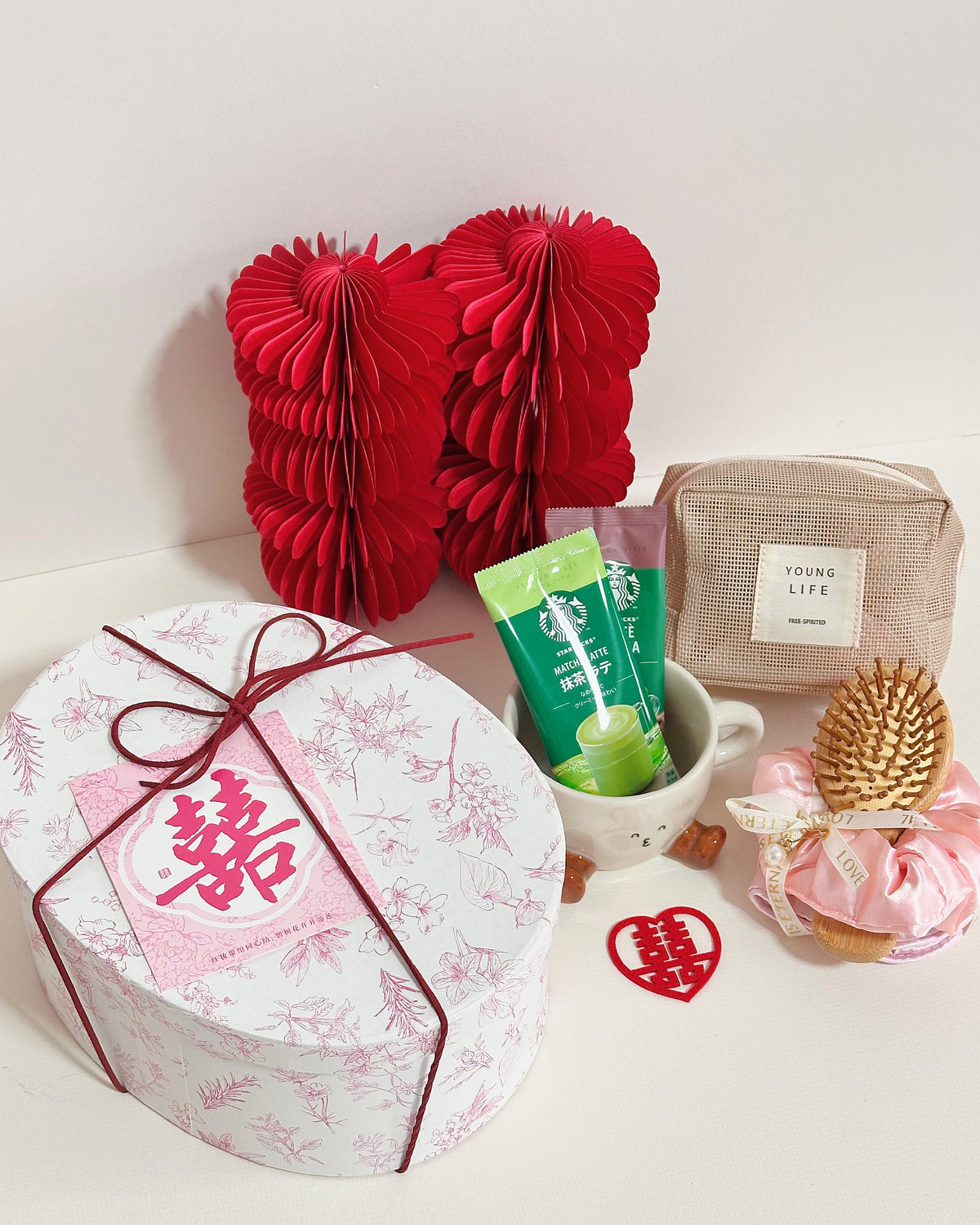 A Valentine's Day themed gift set arranged on a white surface against a white background. The set includes a round white gift box with a pink floral pattern and a red double happiness symbol, tied with a red ribbon; two red paper honeycomb hearts; a small beige pouch labeled 'YOUNG LIFE'; a white mug containing green and pink tubes; a pink wrapped item with a gold pinecone decoration; and a small red heart with a double happiness symbol.