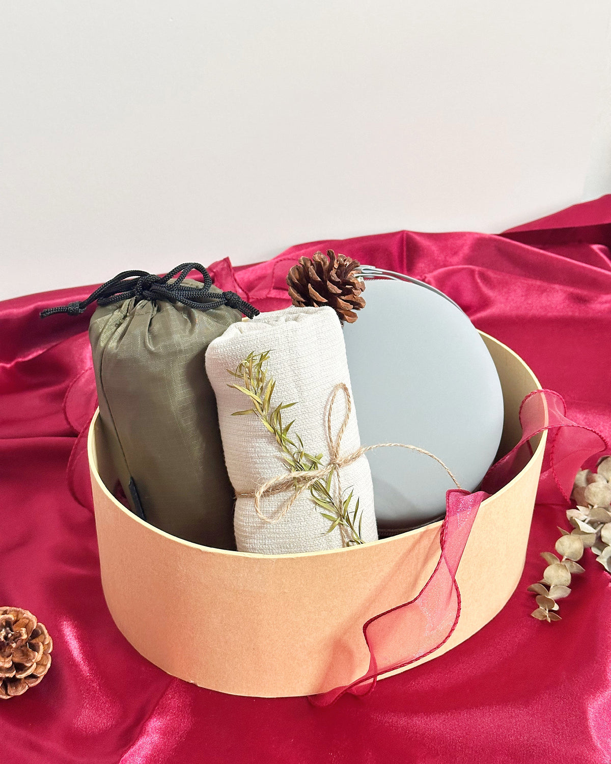A round kraft paper gift box filled with spa-like items, including a sage green drawstring pouch foldable travel duffle bag, a rolled cream linen towel item tied with twine and decorated with a sprig of greenery, a light blue oval object foldable travel neck pillow, and a small pinecone, set on bright pink satin fabric.