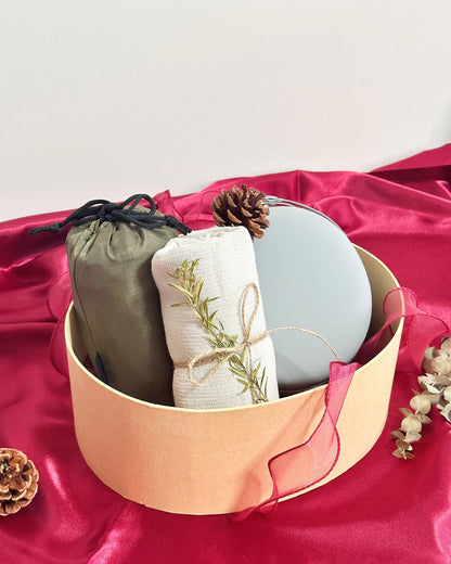 A round kraft paper gift box filled with spa-like items, including a sage green drawstring pouch foldable travel duffle bag, a rolled cream linen towel item tied with twine and decorated with a sprig of greenery, a light blue oval object foldable travel neck pillow, and a small pinecone, set on bright pink satin fabric.